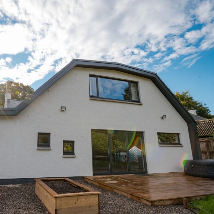 Modern white house with large windows, a wooden deck, and raised garden beds in the backyard.