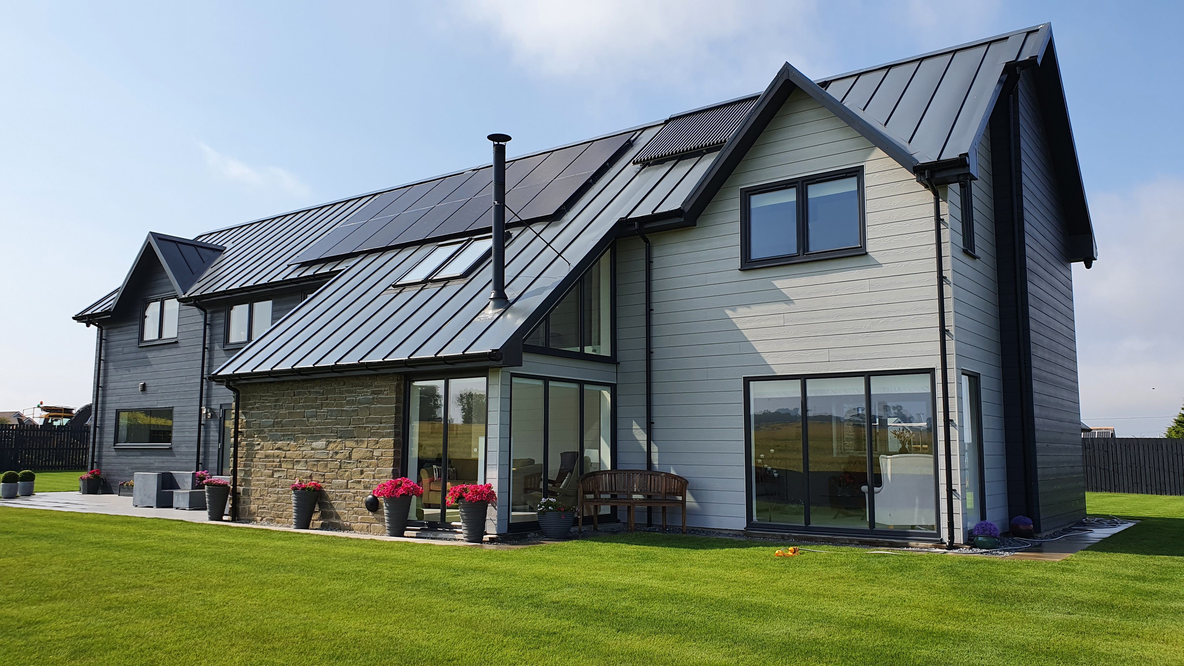 Modern detached house with large windows, metal roof, and solar panels on the roof, surrounded by a well-kept green lawn
