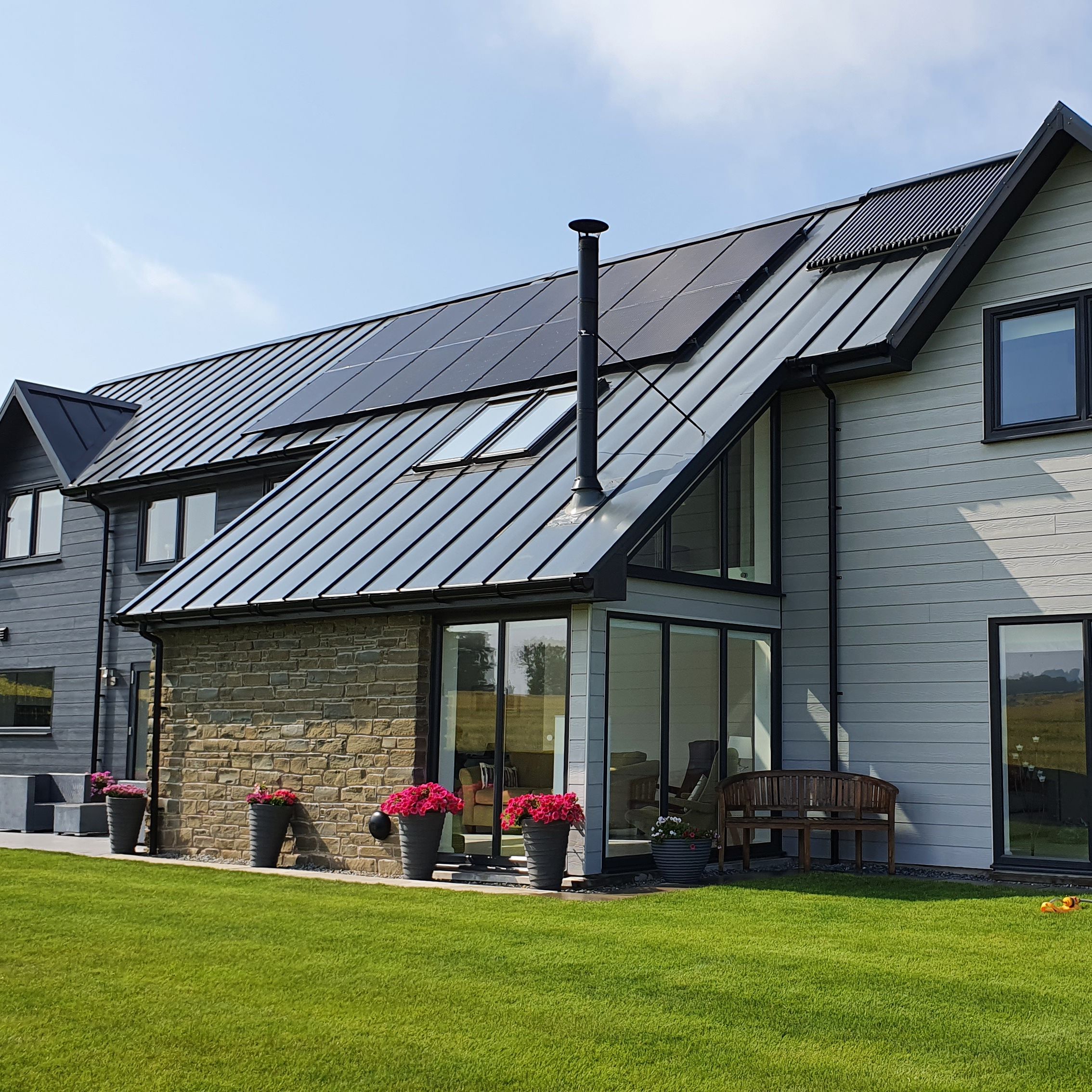 Modern detached house with large windows, metal roof, and solar panels on the roof, surrounded by a well-kept green lawn