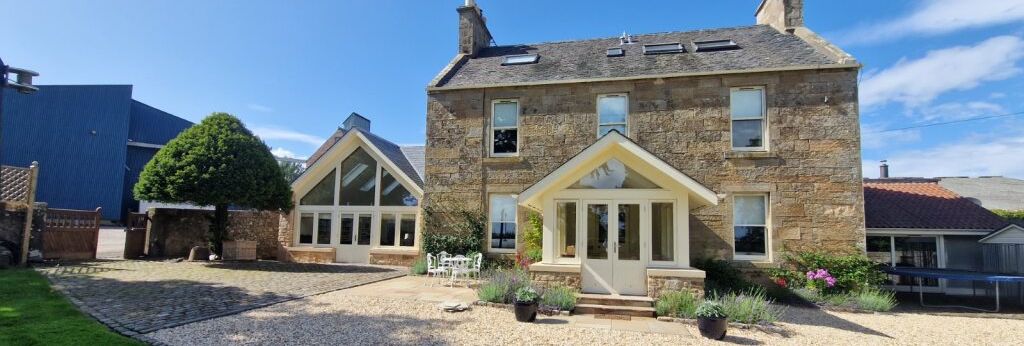 Stone house with modern glass extension, gravel driveway, green lawn, and blue sky