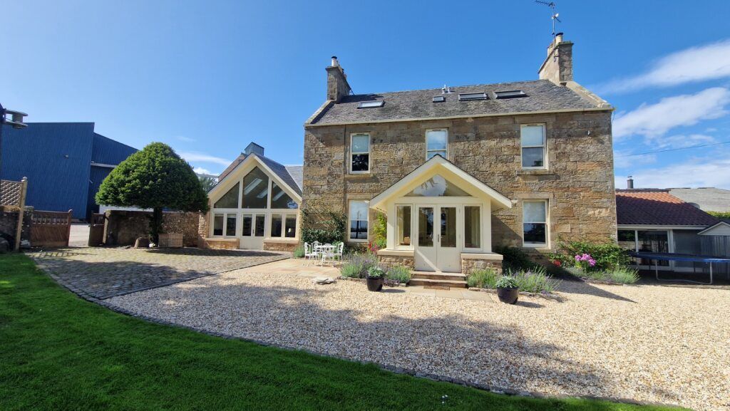 Stone house with modern glass extension, gravel driveway, green lawn, and blue sky
