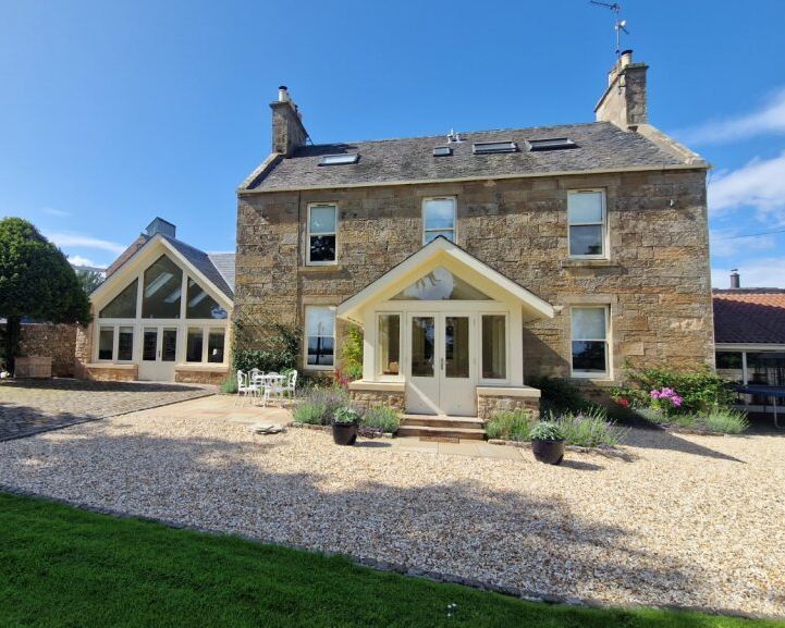 Stone house with modern glass extension, gravel driveway, green lawn, and blue sky