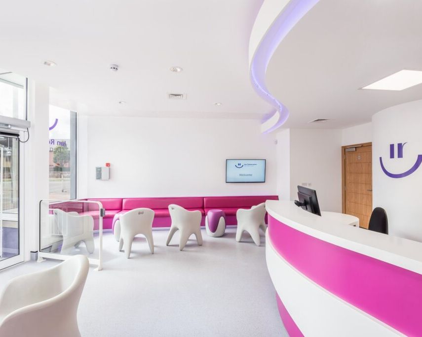 Modern dental or medical clinic waiting area with pink and white decor, curved reception desk, white chairs, and a welcoming atmosphere.