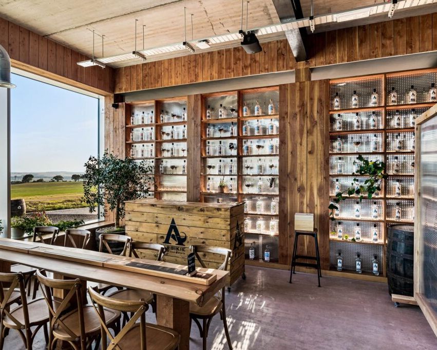 A modern distillery tasting room with wooden interiors, shelves of liquor bottles, large windows, and countryside views.