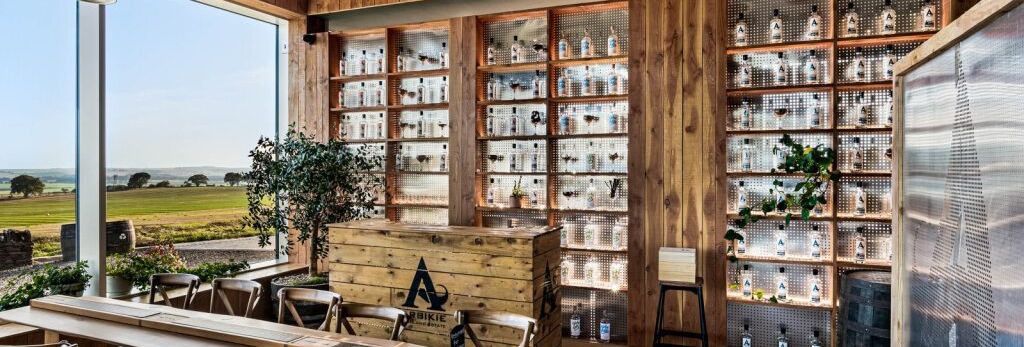 A modern distillery tasting room with wooden interiors, shelves of liquor bottles, large windows, and countryside views.