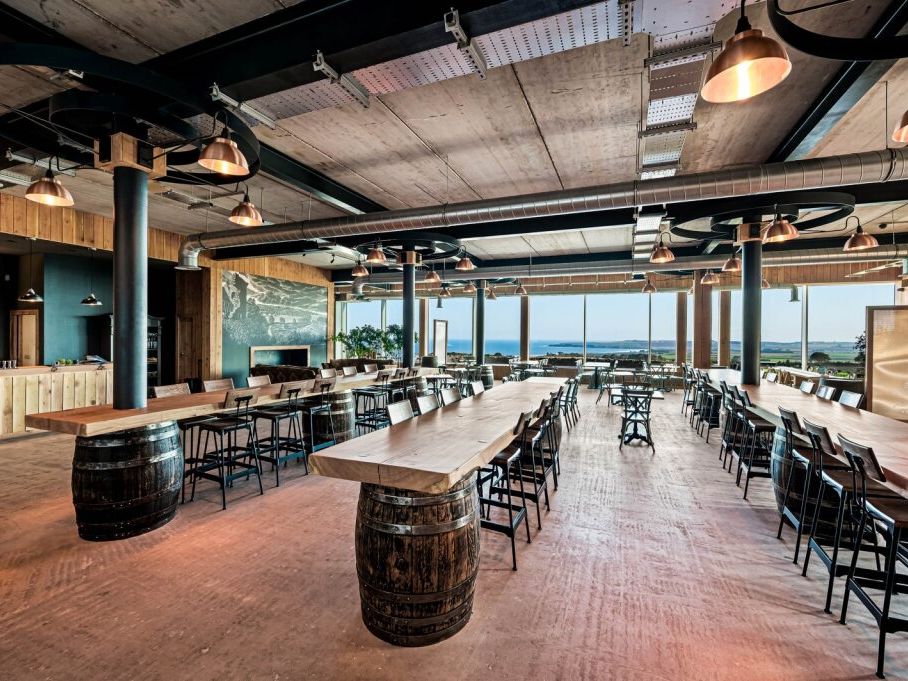 Spacious modern dining area with long wooden tables, barrel bases, industrial lighting, and large windows overlooking a scenic landscape.