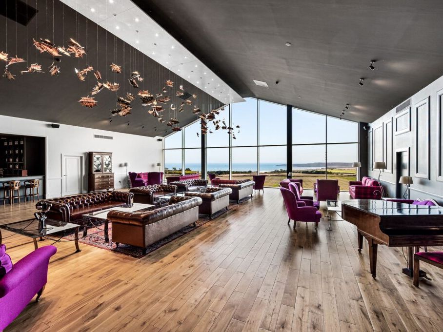 Modern lounge area with large windows overlooking a scenic landscape and ocean, featuring purple armchairs, brown leather sofas, a bar, and decorative ceiling art.