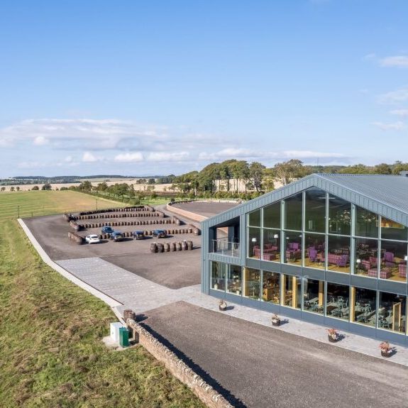 Modern glass-fronted building surrounded by green fields under a blue sky