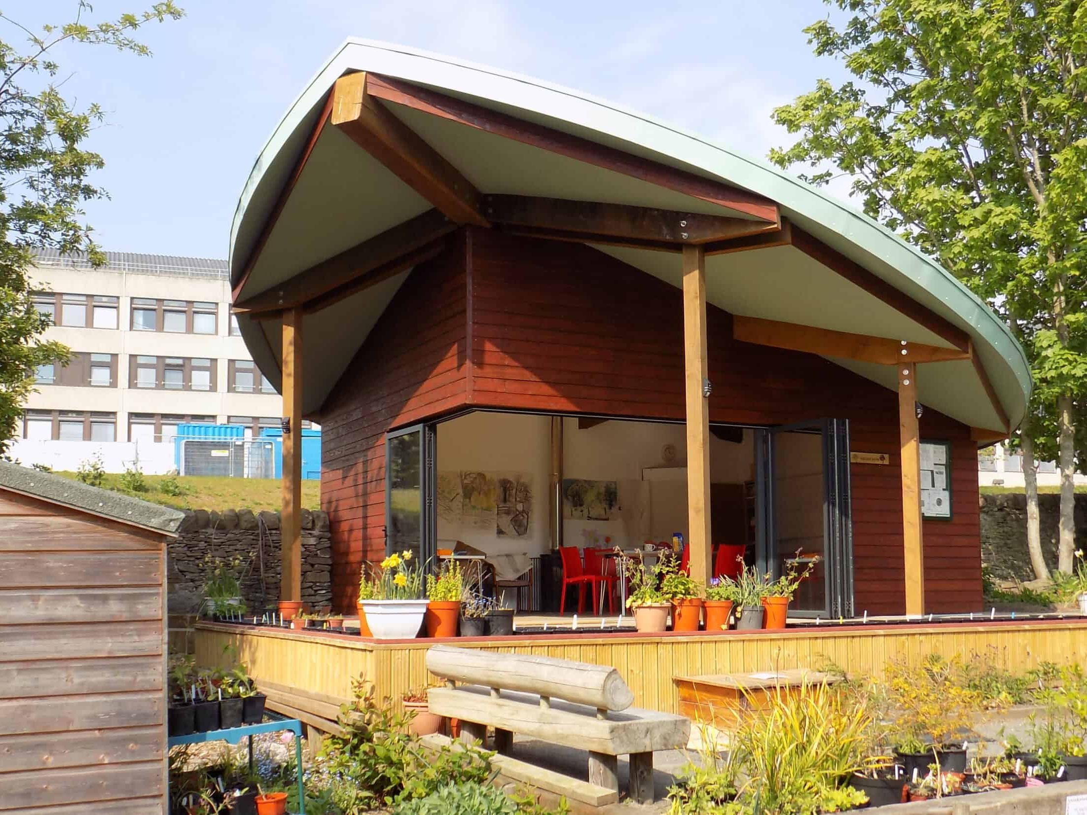 A modern wooden building with a unique curved roof, surrounded by plants and trees.