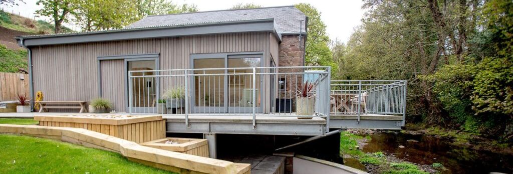 Modern house with a raised patio over a stream, surrounded by landscaped garden and trees