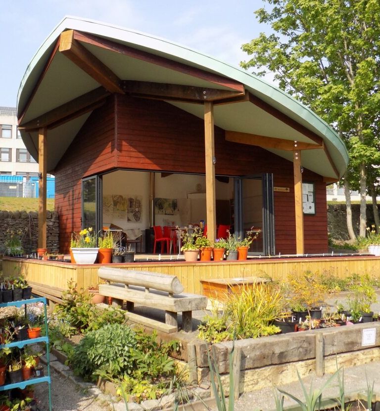 A modern wooden garden building with a curved roof, surrounded by plants and a shed.