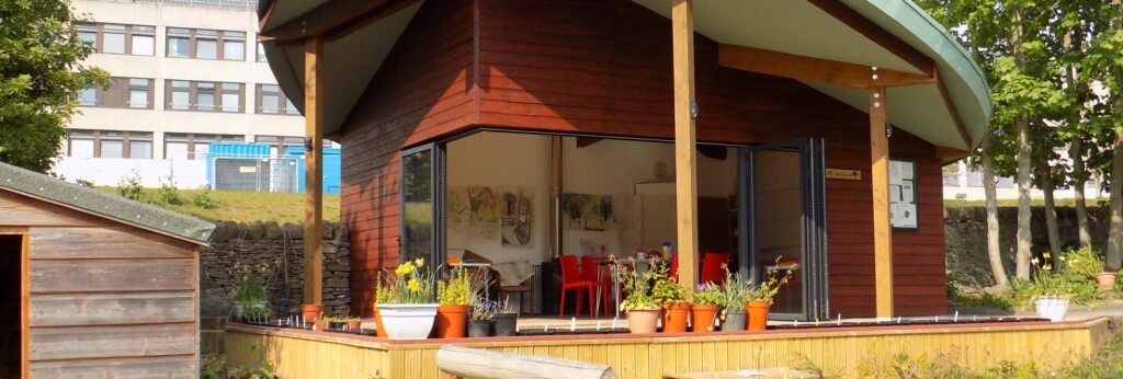 A modern wooden garden building with a curved roof, surrounded by plants and a shed.