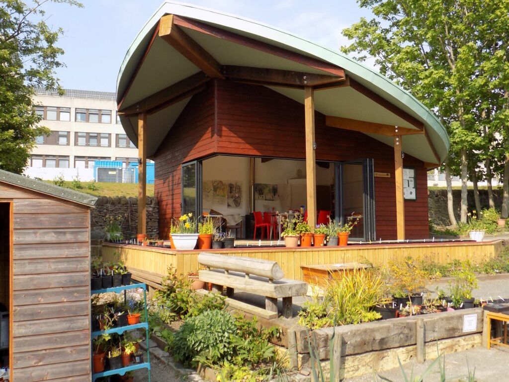 A modern wooden garden building with a curved roof, surrounded by plants and a shed.