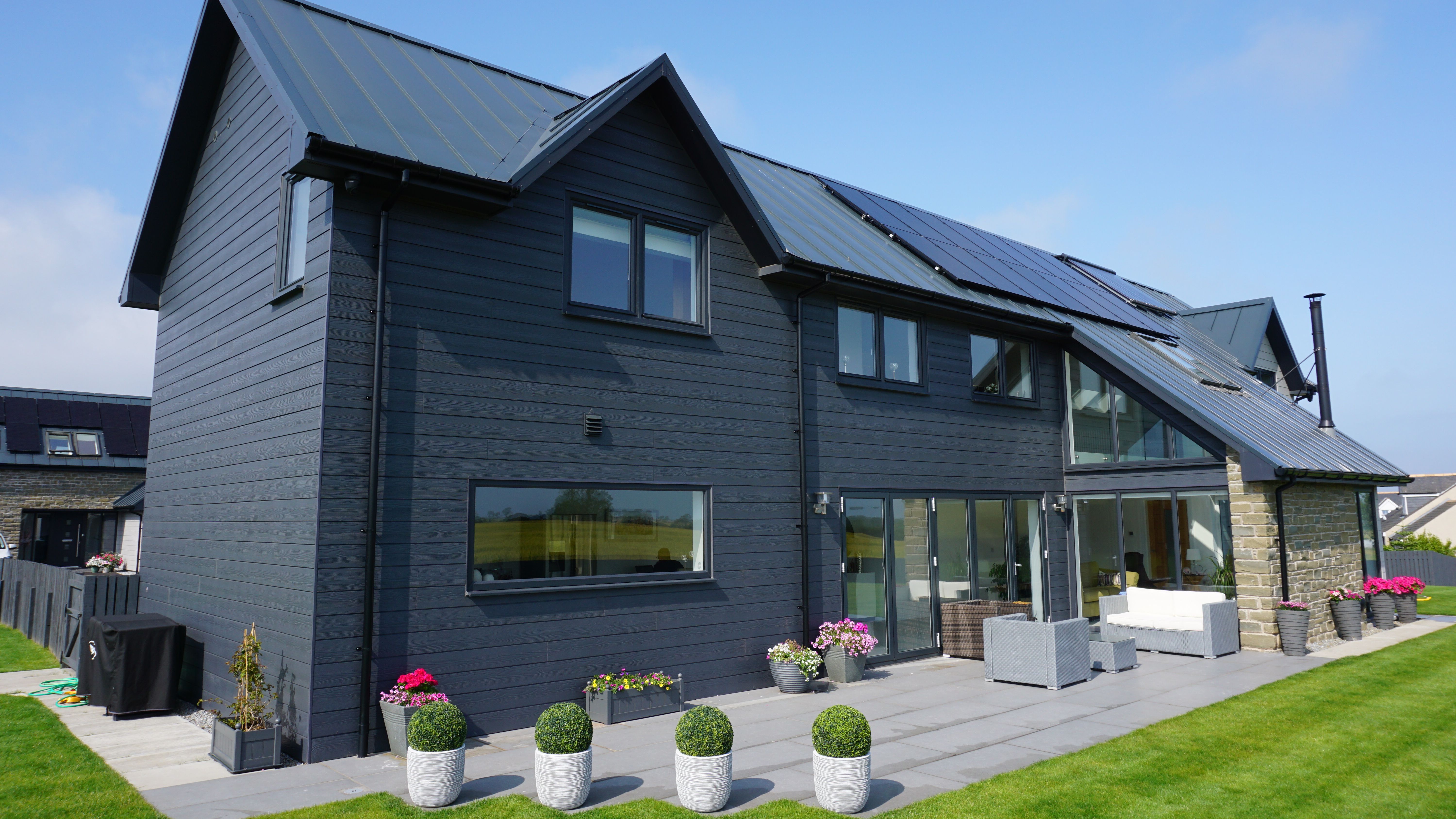 Modern dark grey house with large windows, solar panels on the roof, and a neatly landscaped patio with potted plants.