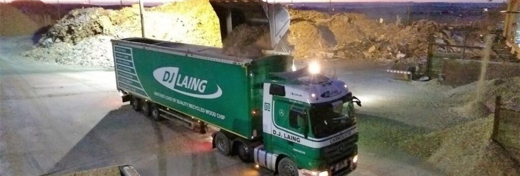 Green DJ Laing truck being loaded with wood chips at an outdoor processing facility during sunset.