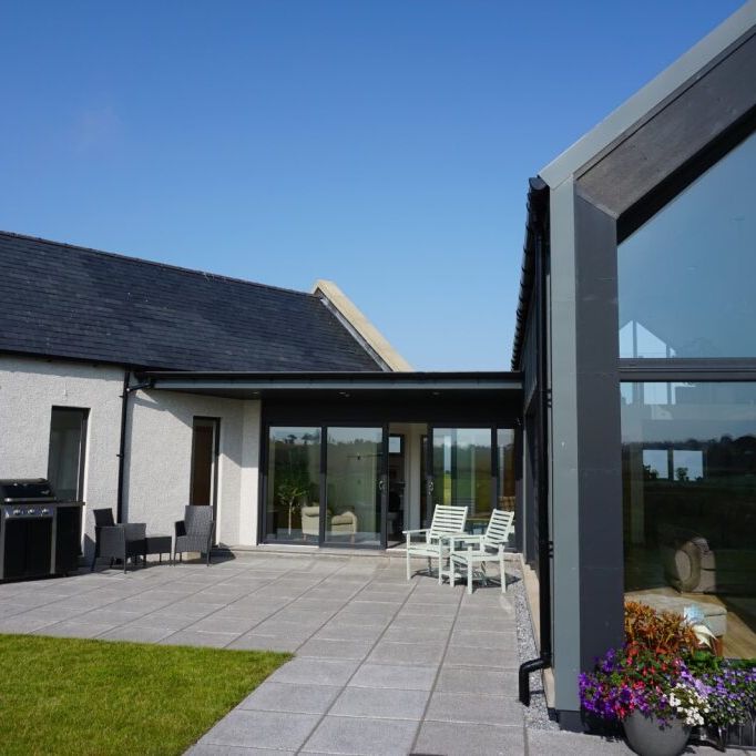 Modern house with large windows, patio seating, and garden under a clear blue sky