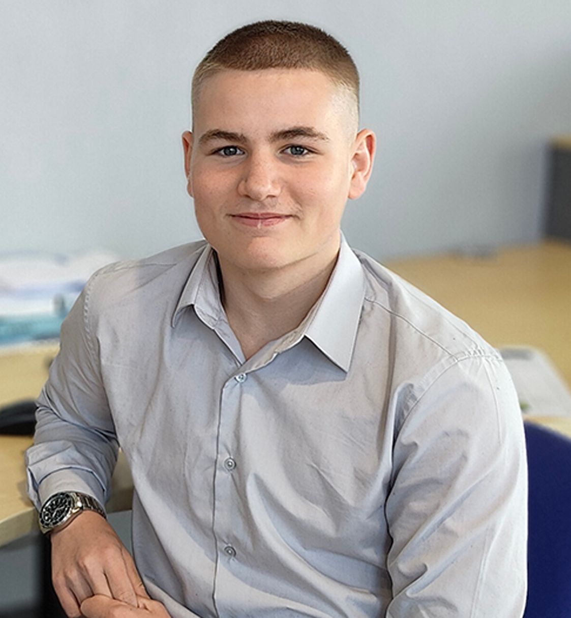 A young man sitting at a desk, wearing a light gray button-up shirt and a wristwatch.