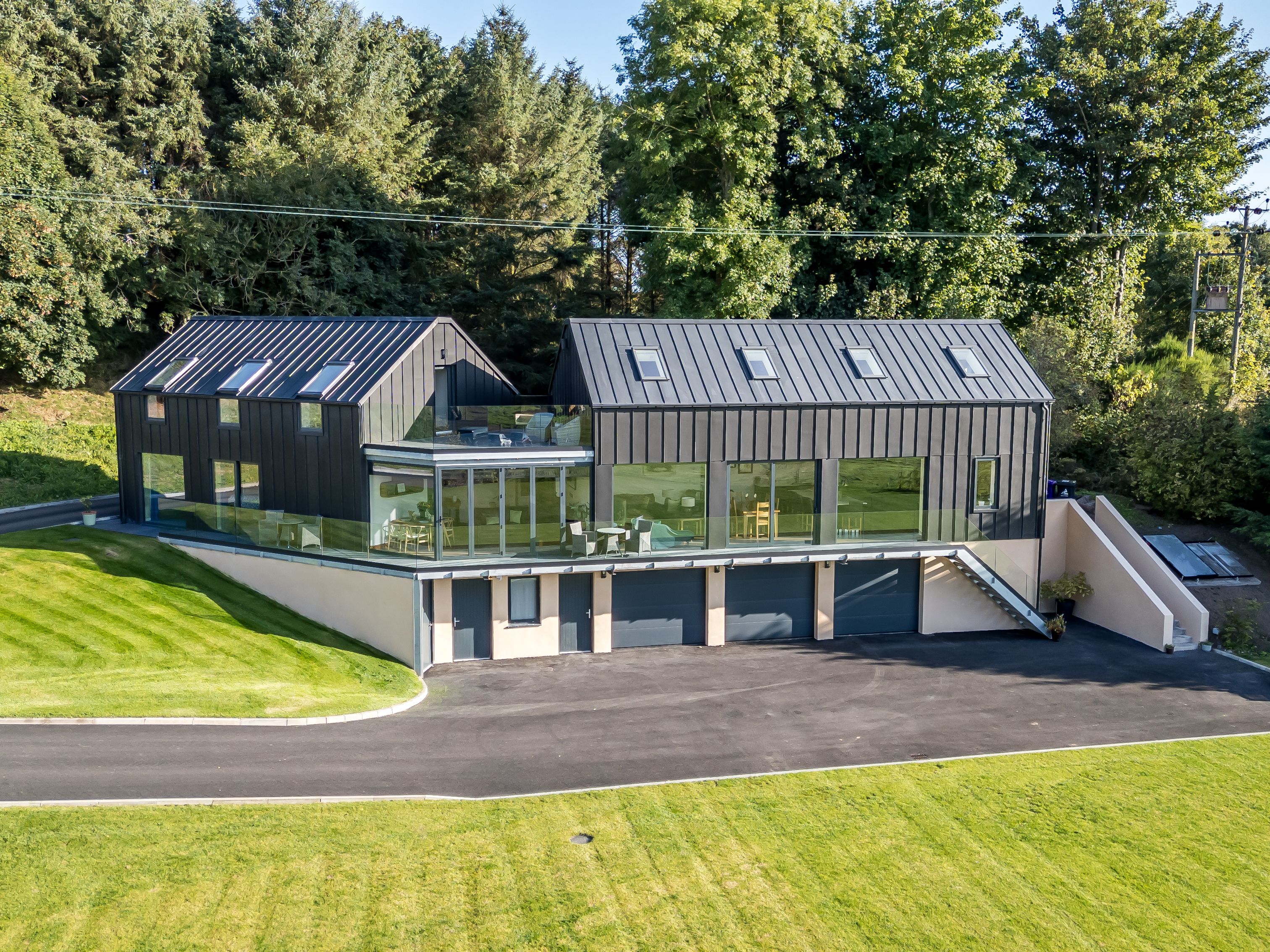 Modern house with black metal roof and large glass windows surrounded by green lawn and trees