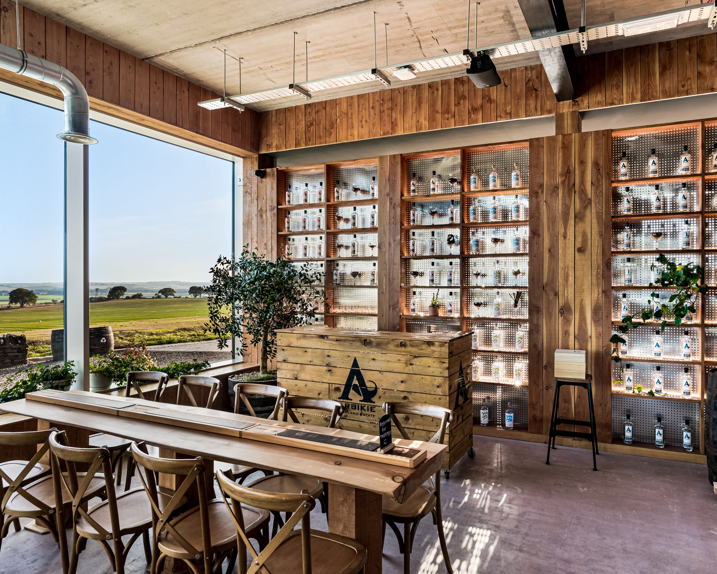 Modern tasting room with wooden decor, large windows overlooking farmland, and a display of bottles on backlit shelves.