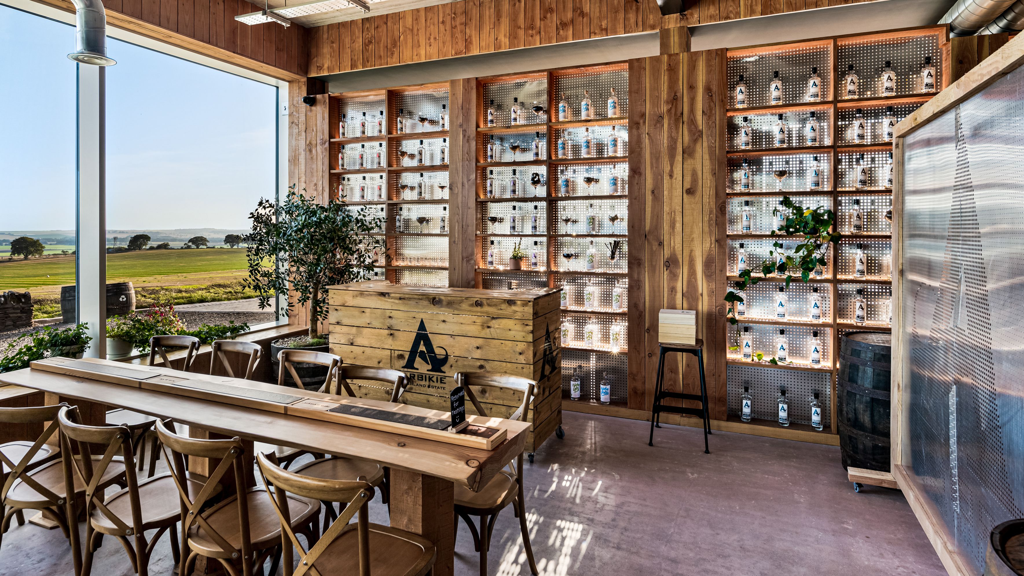 Modern tasting room with wooden decor, large windows overlooking farmland, and a display of bottles on backlit shelves.