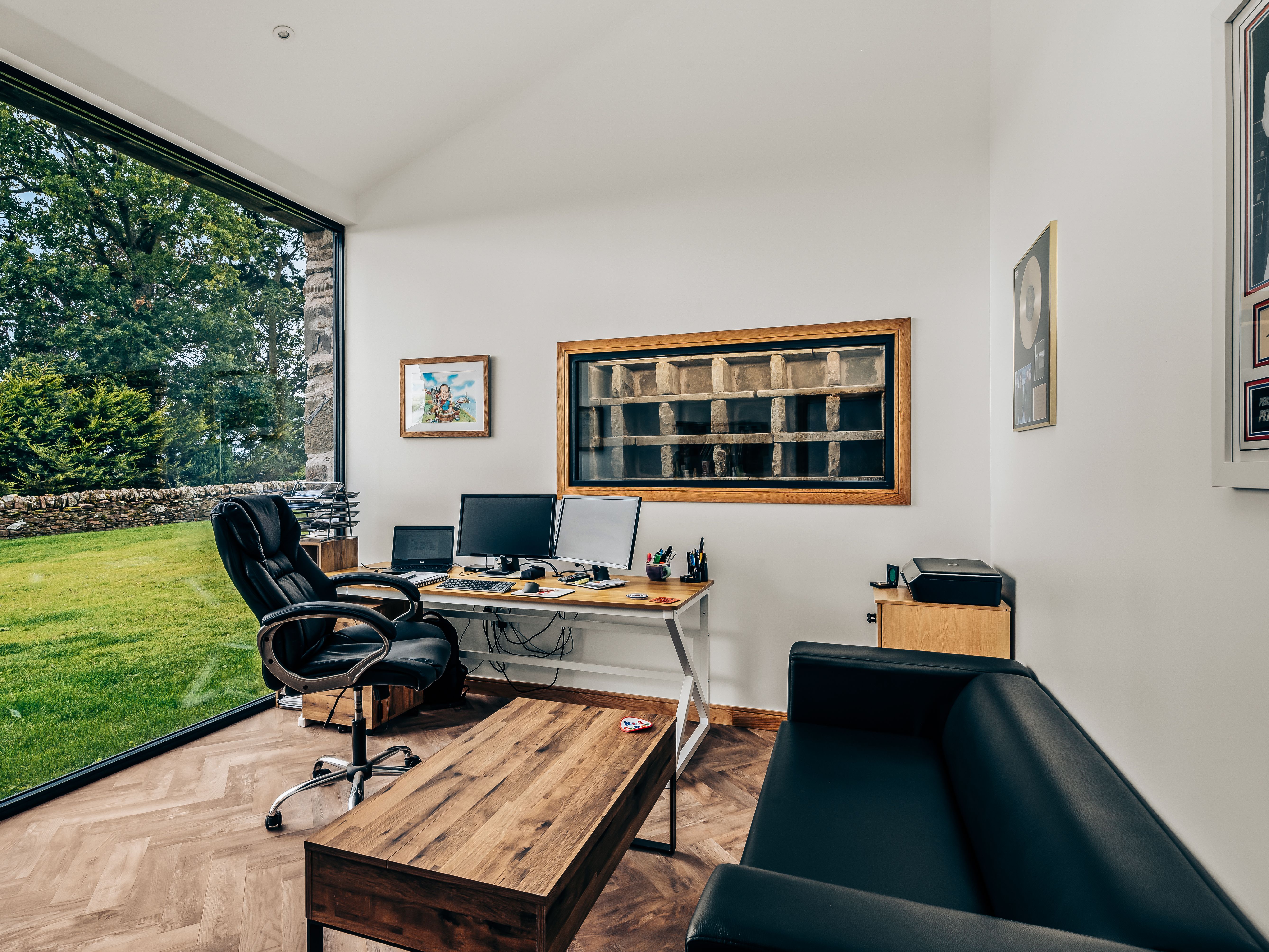 Modern office space with large window overlooking a garden, black leather chair and couch, wooden desk, and multiple computer monitors.