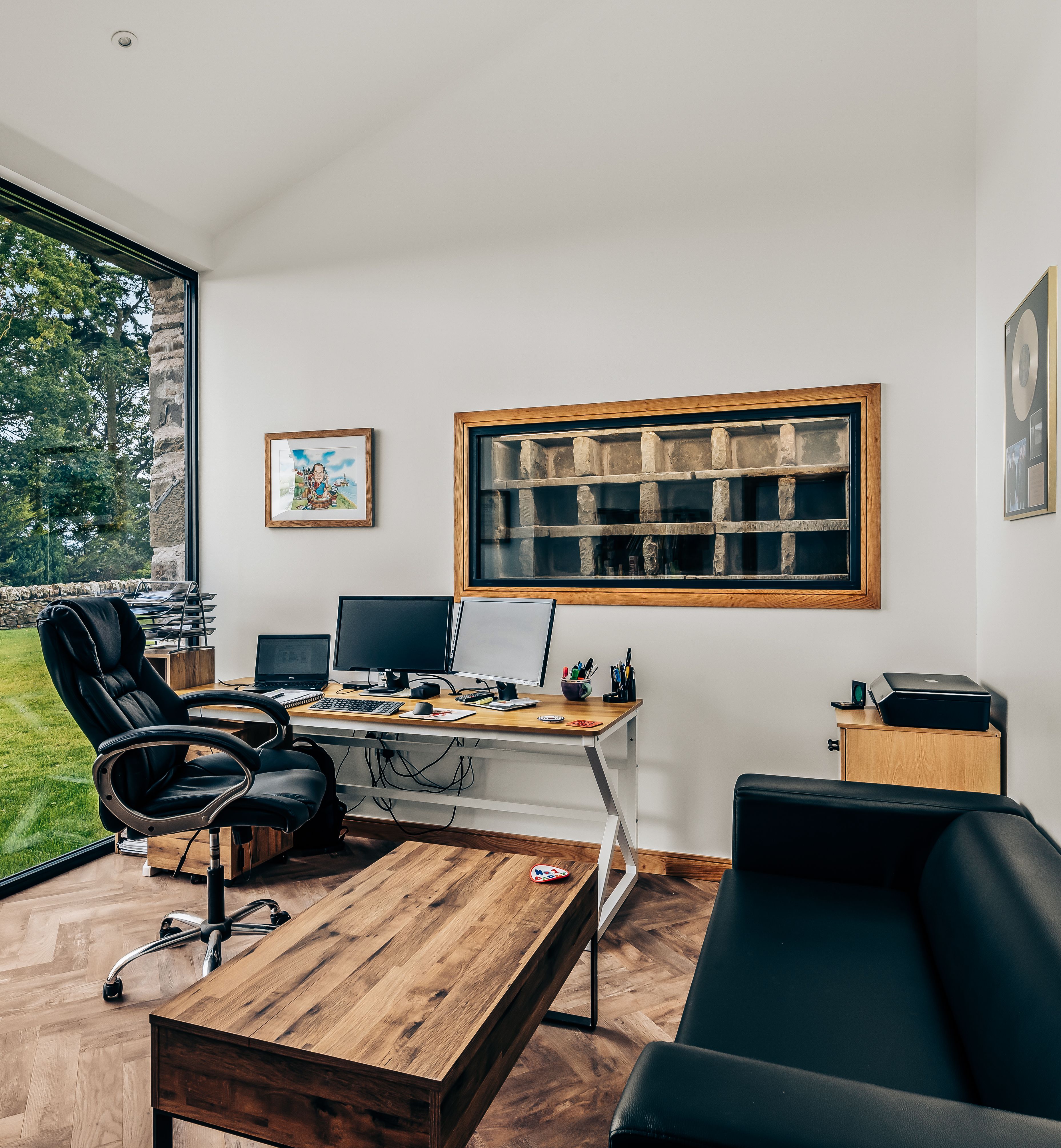 Modern office space with large window overlooking a garden, black leather chair and couch, wooden desk, and multiple computer monitors.