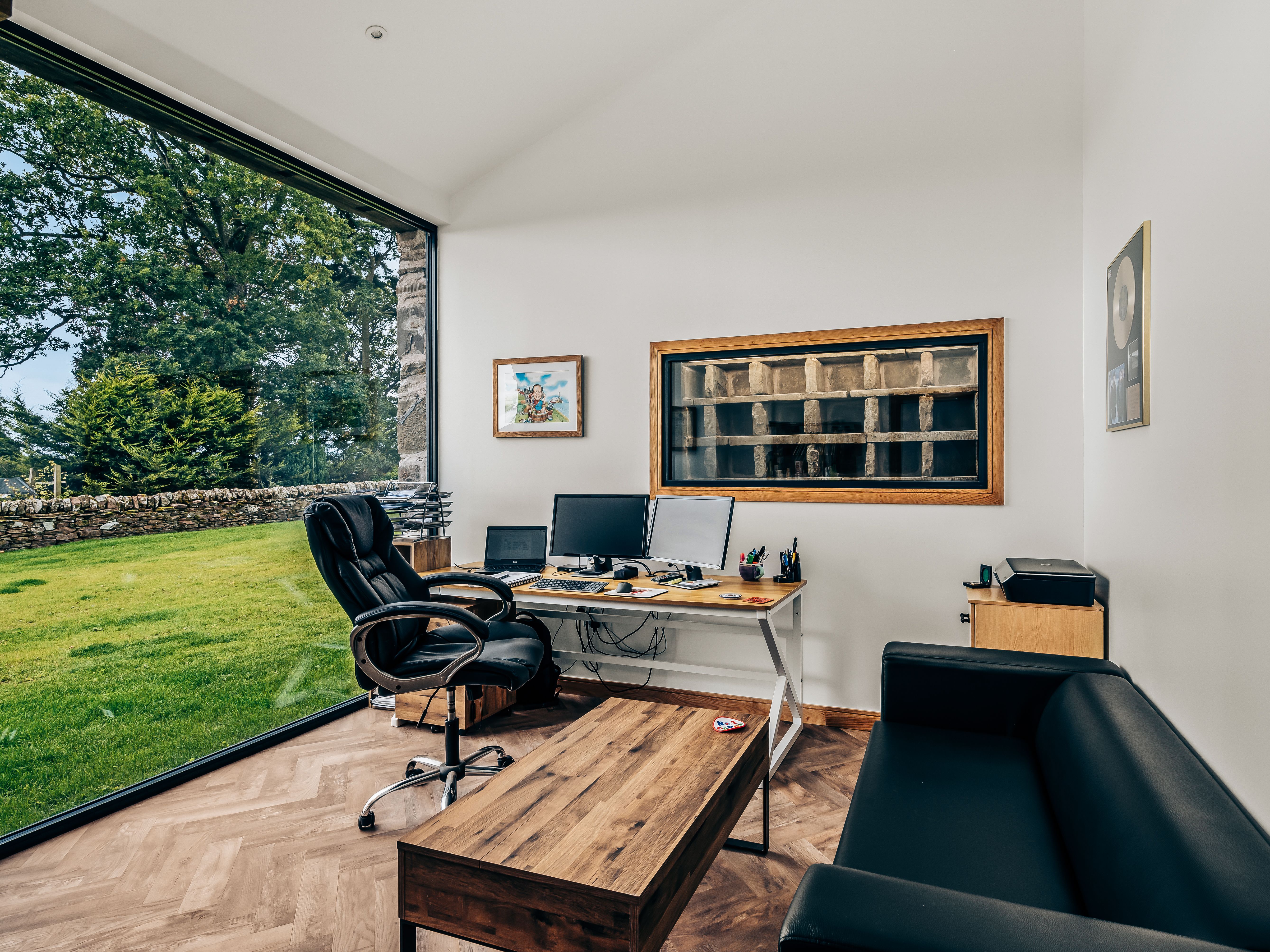 Modern office space with large window overlooking a garden, black leather chair and couch, wooden desk, and multiple computer monitors.