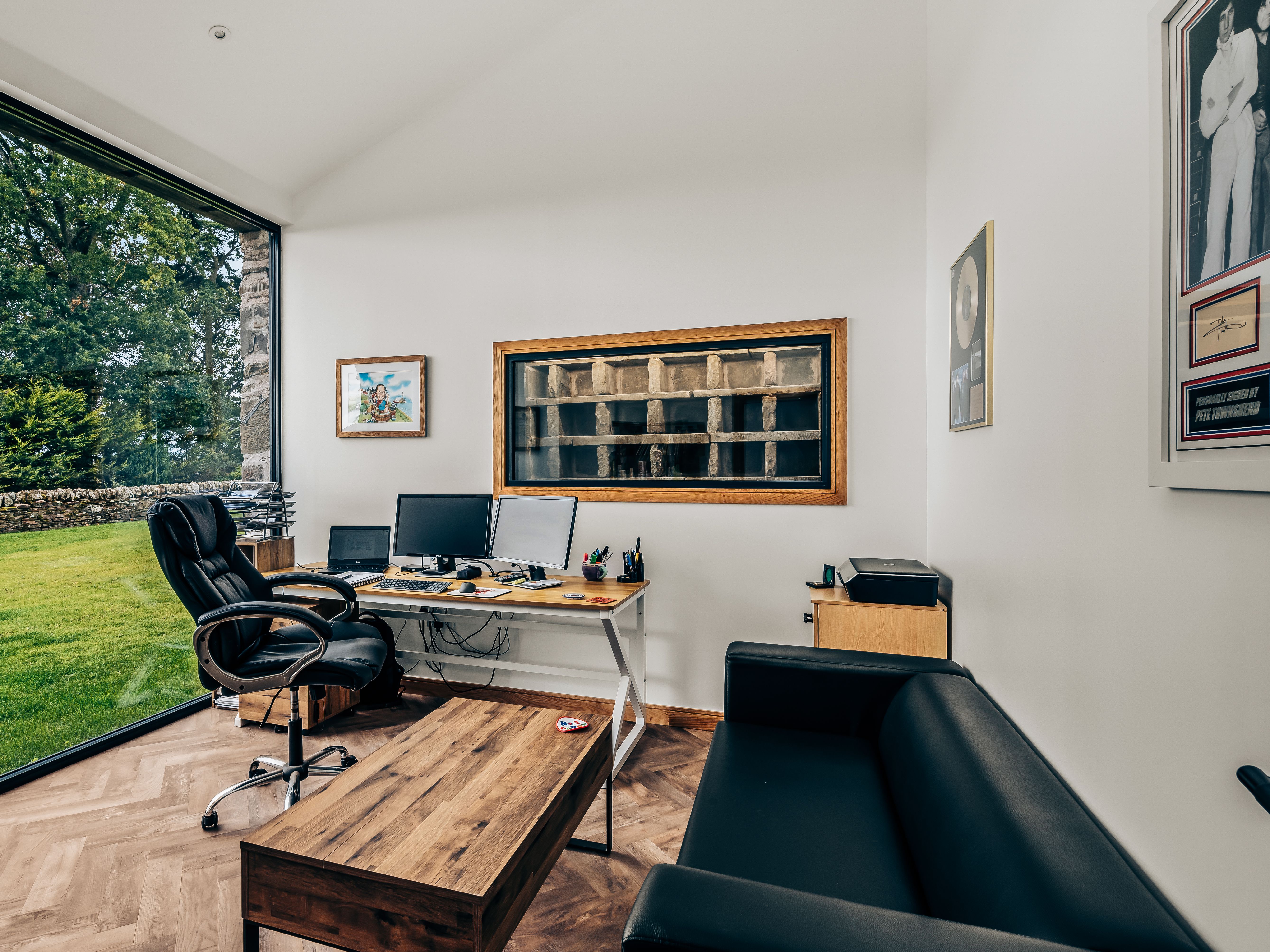 Modern office space with large window overlooking a garden, black leather chair and couch, wooden desk, and multiple computer monitors.