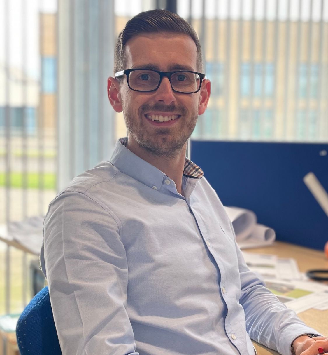 Smiling man with glasses sitting at an office desk with papers and a notebook.