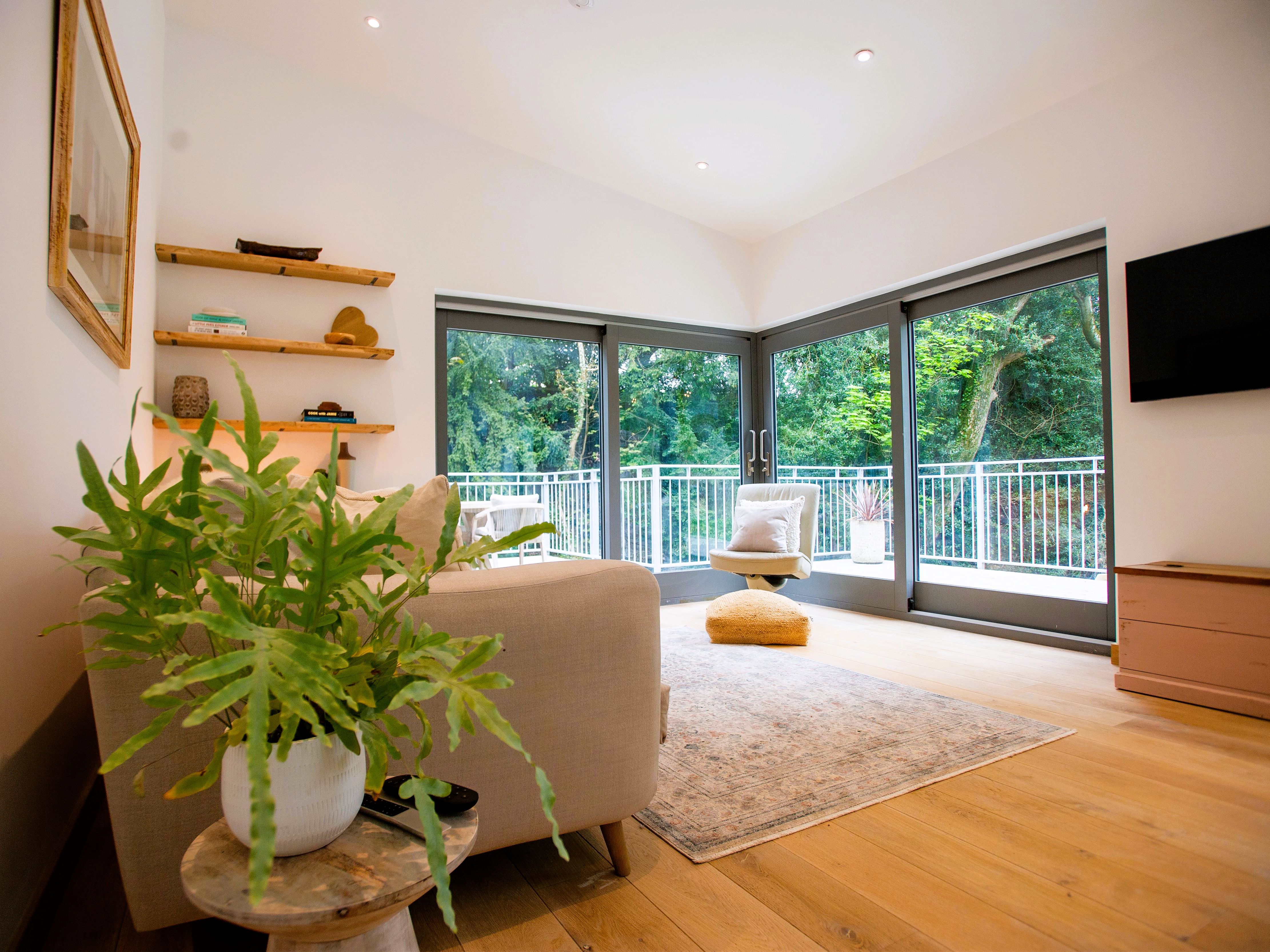Modern living room with large windows, a beige sofa, indoor plants, and a view of greenery outside.