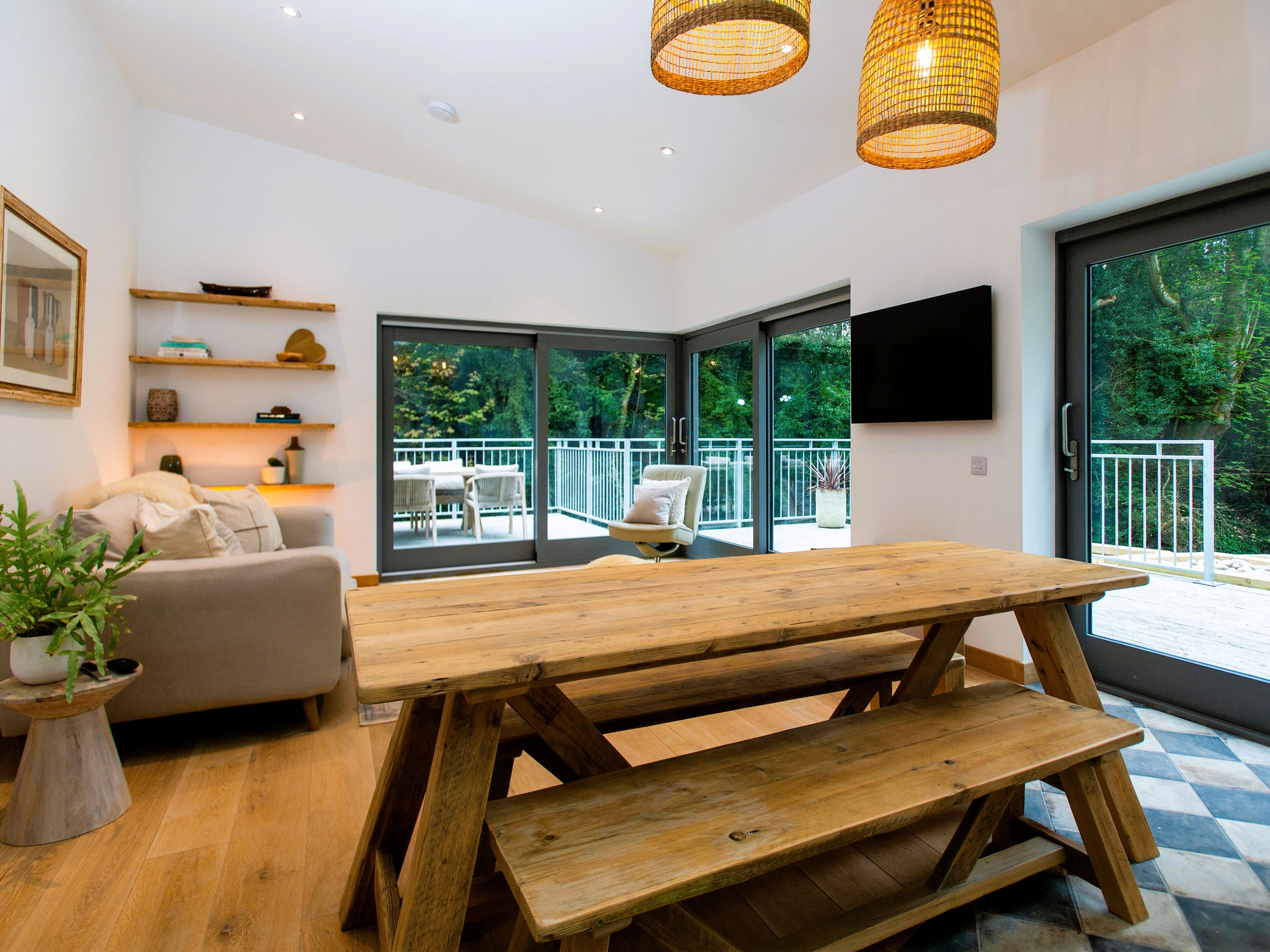 Bright, modern living and dining area with rustic wooden table and bench, large windows, and view of balcony and trees.