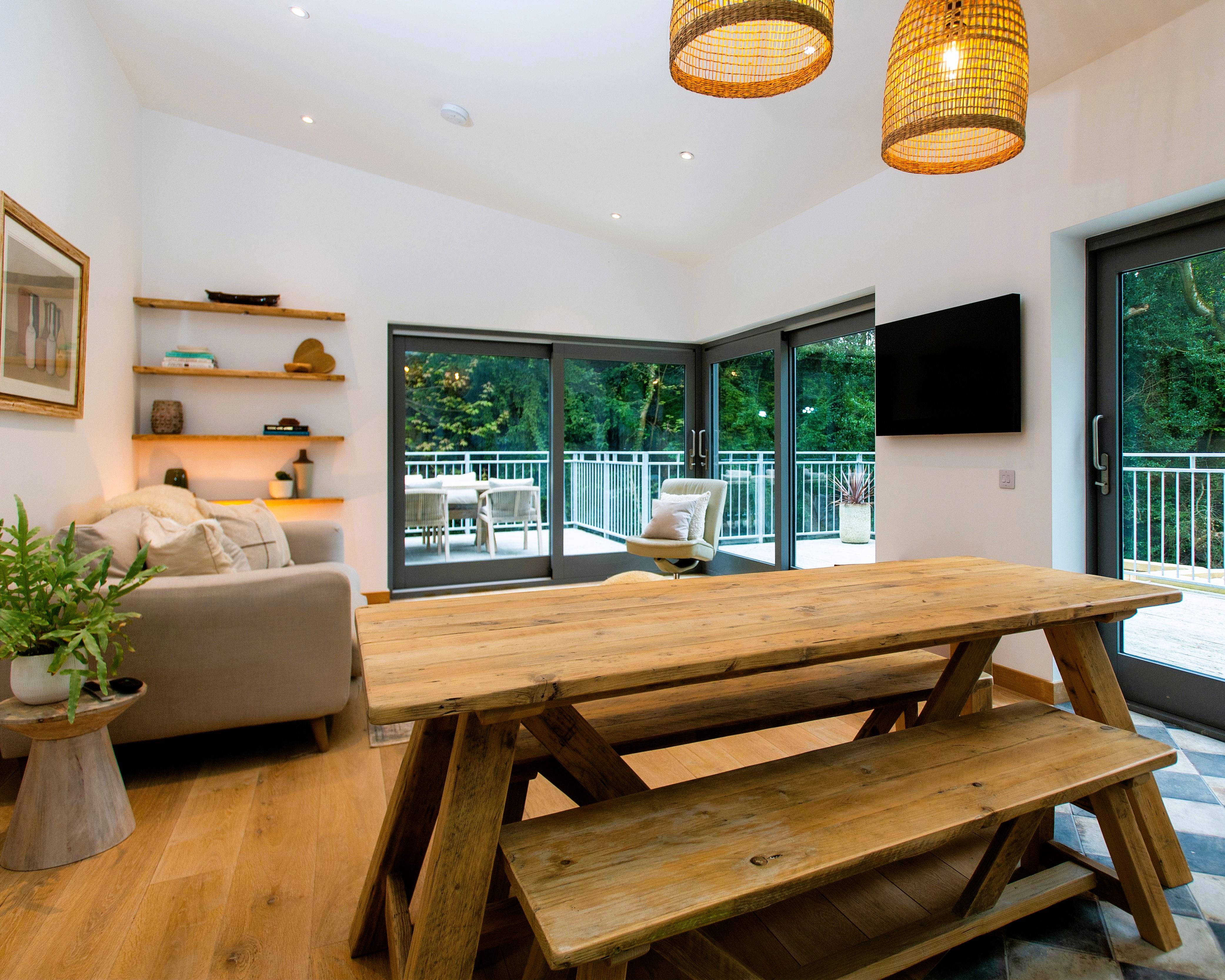 Bright, modern living and dining area with rustic wooden table and bench, large windows, and view of balcony and trees.