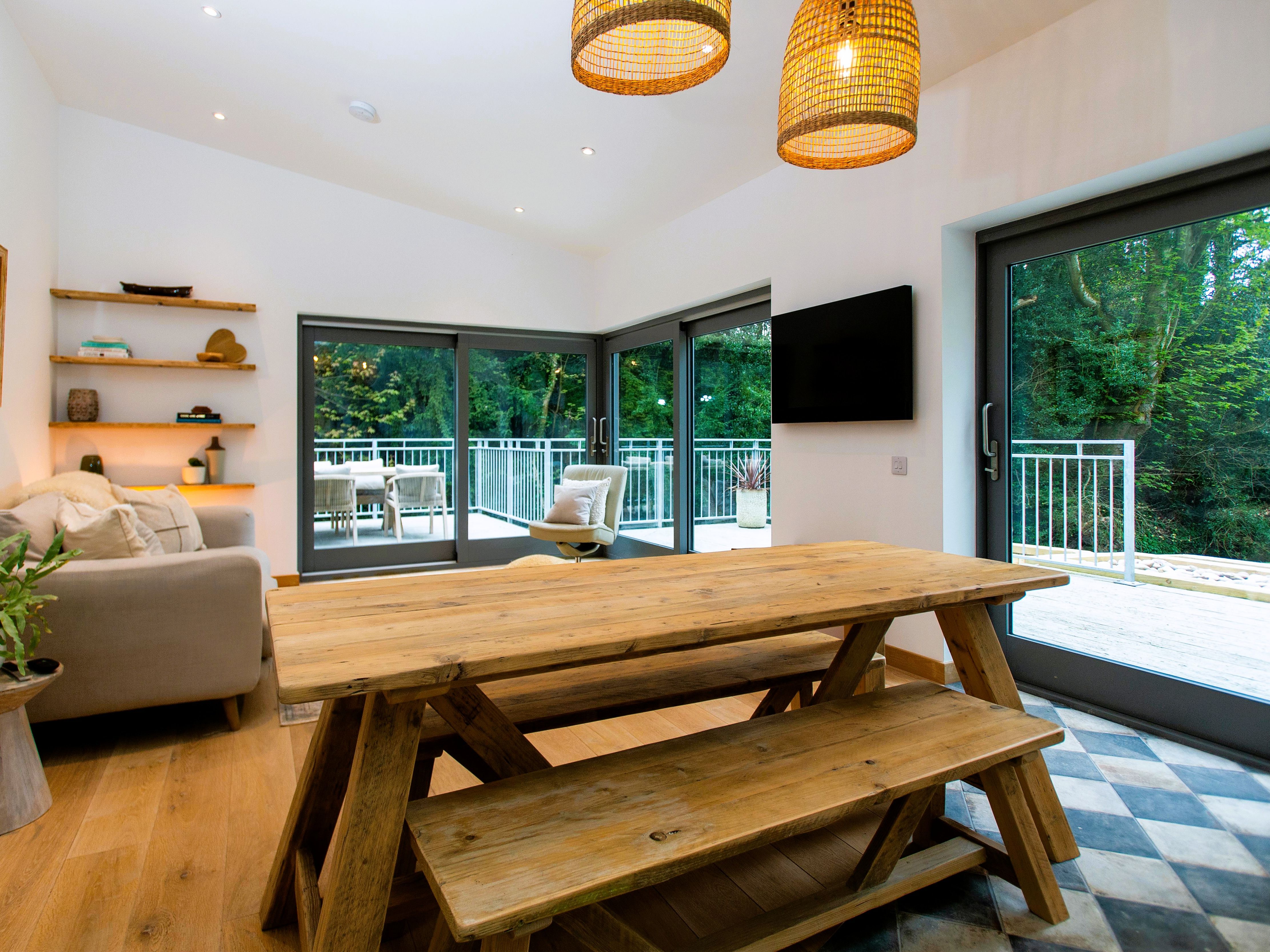 Bright, modern living and dining area with rustic wooden table and bench, large windows, and view of balcony and trees.