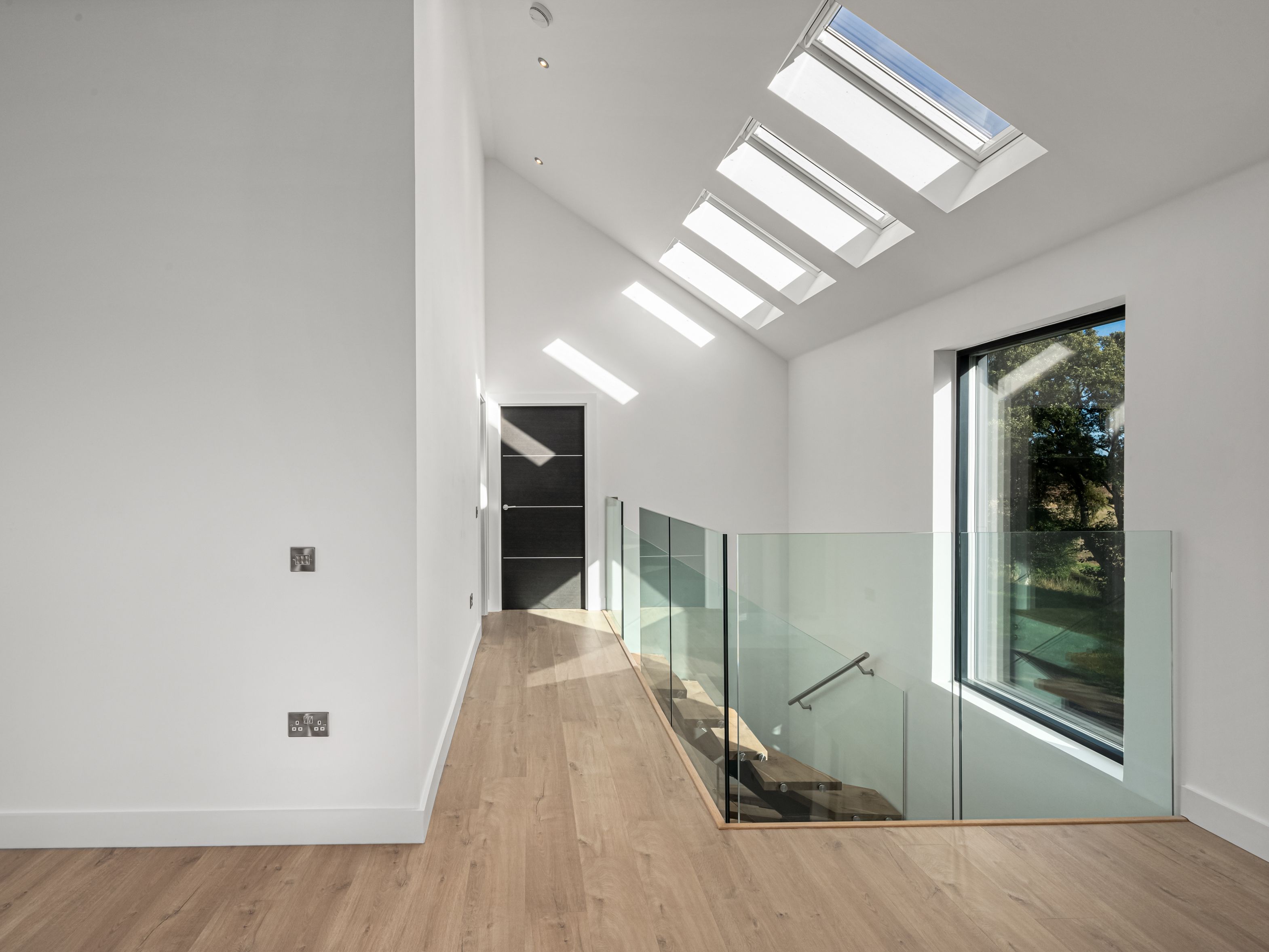 Modern hallway with wooden floor, glass stair railing, large window, and skylights