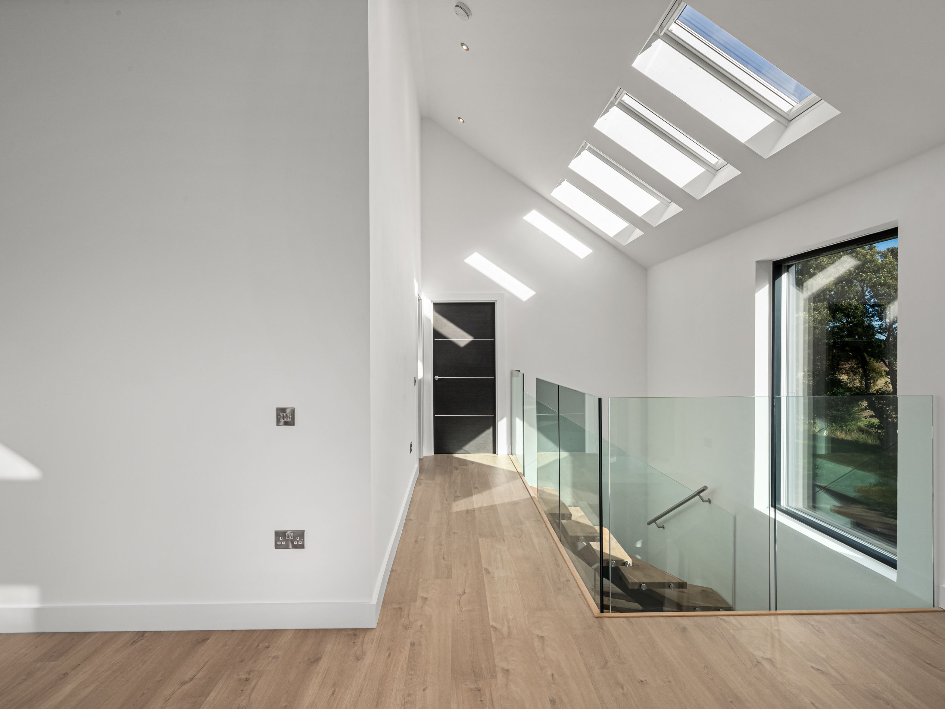 Modern hallway with wooden floor, glass stair railing, large window, and skylights