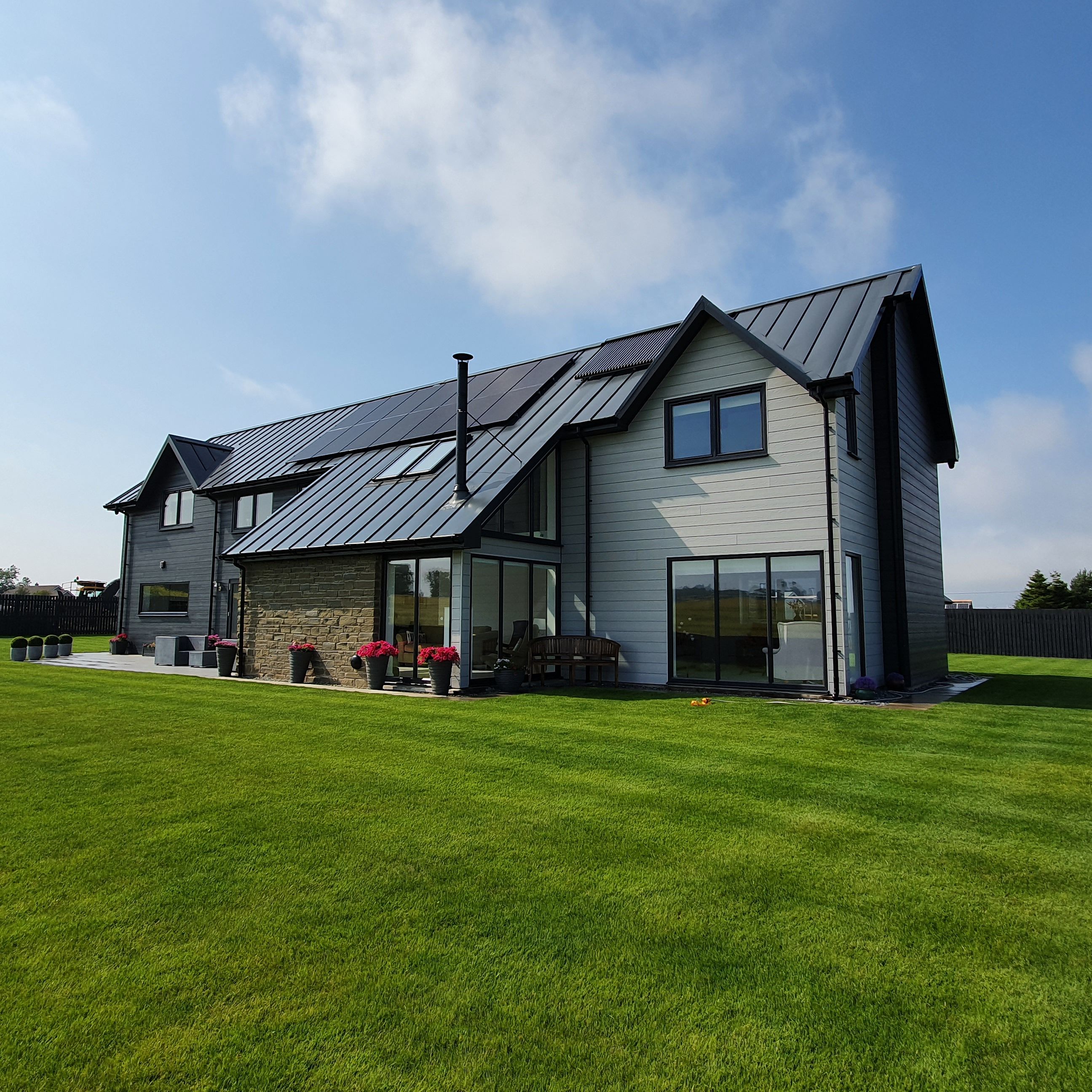 Modern two-story house with large windows, metal roof, and spacious green lawn under a blue sky.
