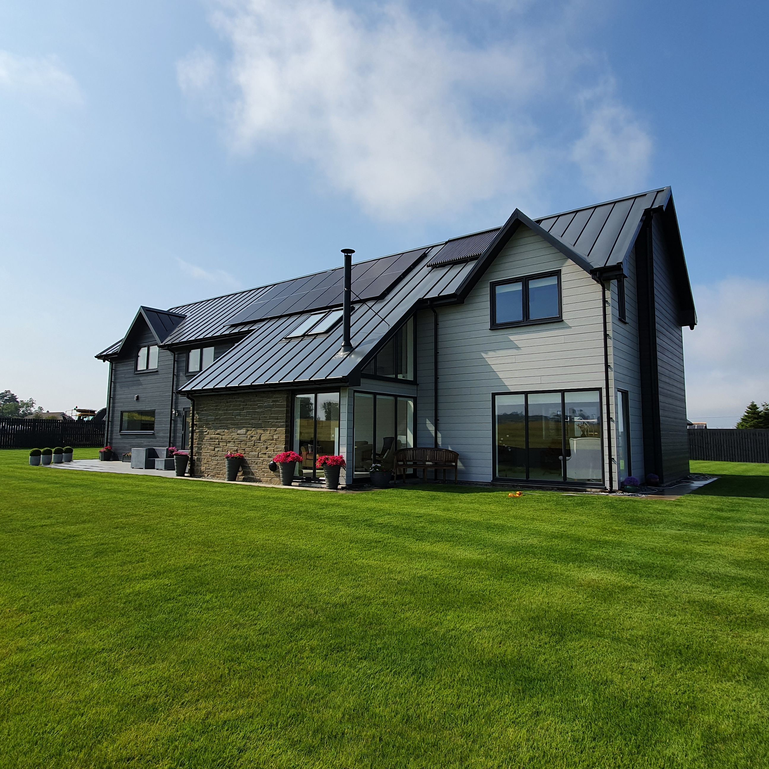 Modern two-story house with large windows, metal roof, and spacious green lawn under a blue sky.