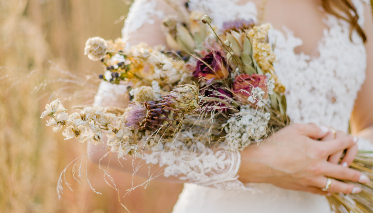 A bride in a lace wedding dress holding a rustic bouquet of dried flowers and grasses, featuring soft hues of cream, purple, and pink, set against a softly blurred natural background.