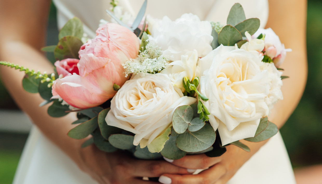 A bride in a white dress holding a bouquet of soft pink and ivory roses, white flowers, and eucalyptus leaves, arranged in a natural style, with her hands gently clasping the stems.