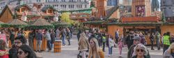 A festive outdoor market with wooden stalls, Christmas decorations, and a large windmill structure. Crowds of people walk around, enjoying the lively atmosphere under a blue sky.