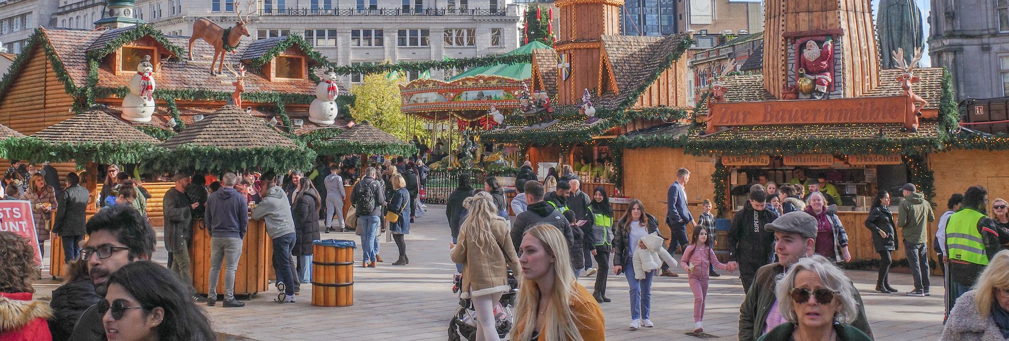 A festive outdoor market with wooden stalls, Christmas decorations, and a large windmill structure. Crowds of people walk around, enjoying the lively atmosphere under a blue sky.