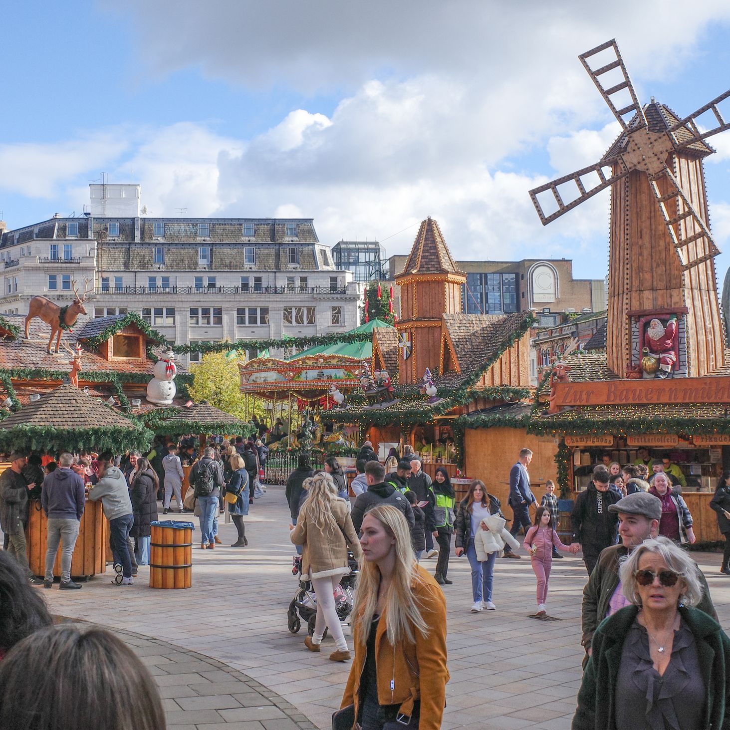 A festive outdoor market with wooden stalls, Christmas decorations, and a large windmill structure. Crowds of people walk around, enjoying the lively atmosphere under a blue sky.