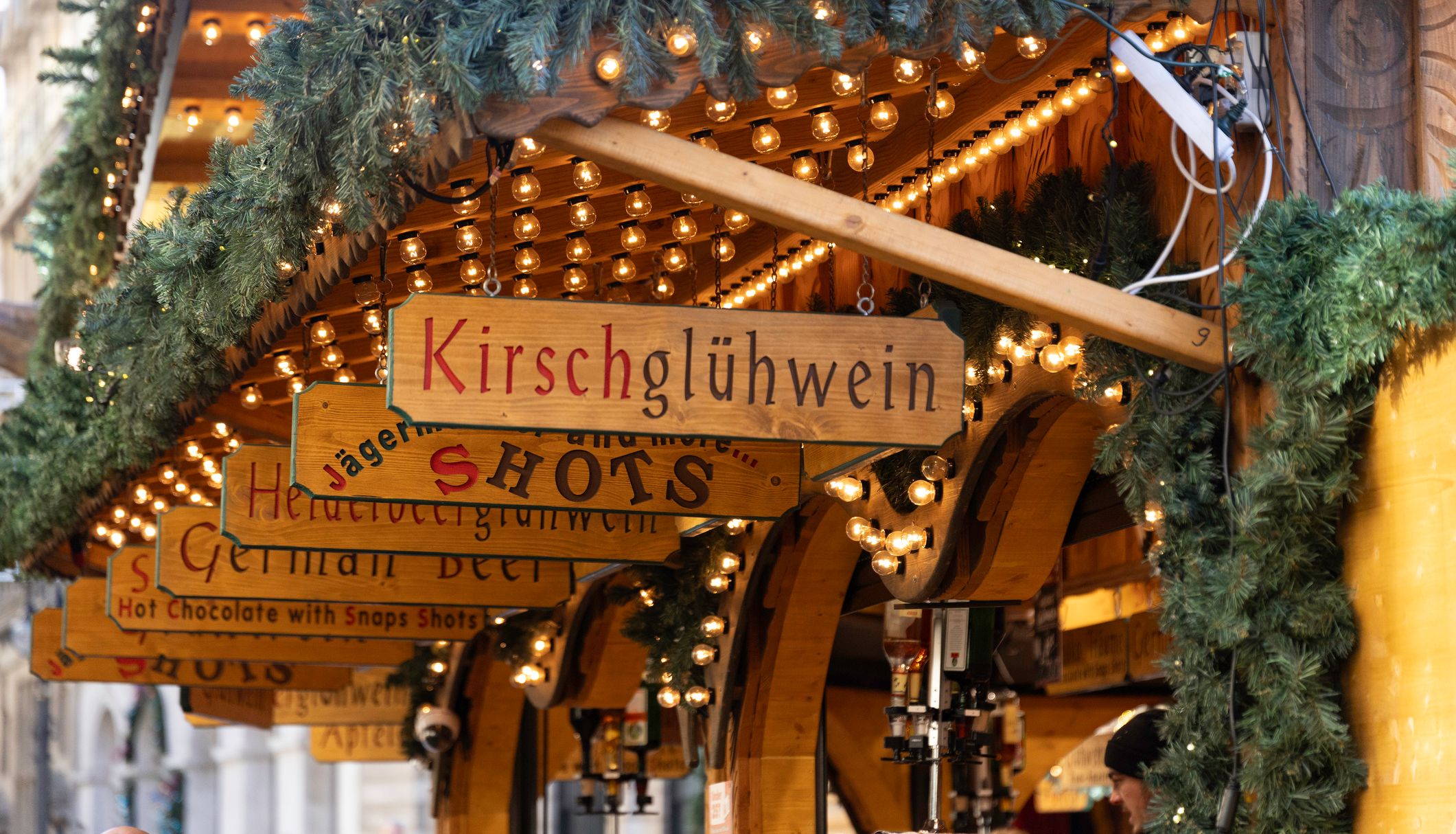 A close-up of a festive market stall decorated with string lights and pine garlands. Wooden signs display