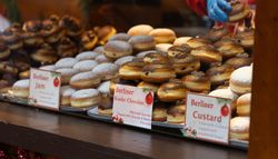 A bakery display with trays of assorted Berliner donuts, including jam, Kinder chocolate, and custard varieties. Each donut is topped with powdered sugar or chocolate.