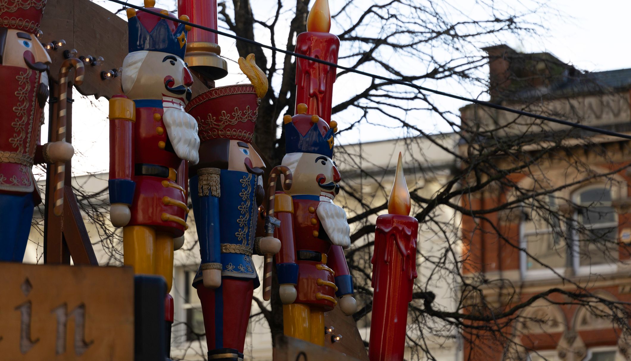 A row of large decorative nutcrackers and red candles displayed outdoors. Bare tree branches and the facade of a historic building are visible in the background.