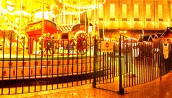 A brightly lit carousel glows in the night, surrounded by a black metal fence with a 