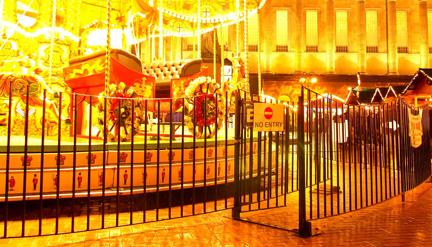 A brightly lit carousel glows in the night, surrounded by a black metal fence with a