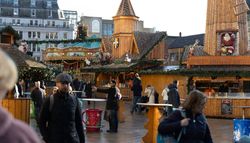 A festive market with wooden stalls, a windmill, and holiday decorations. People walk and take photos, while a carousel and Santa figure add to the Christmas atmosphere.