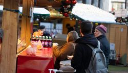 People stand at a festive market stall decorated with ornaments and garlands. Bottles of drinks and small pig figurines are displayed on the counter as they order food.