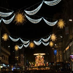 A festive street in Birmingham adorned with glowing Christmas lights, including golden snowflakes and draped garlands, with a vibrant holiday market in the background buzzing with activity.